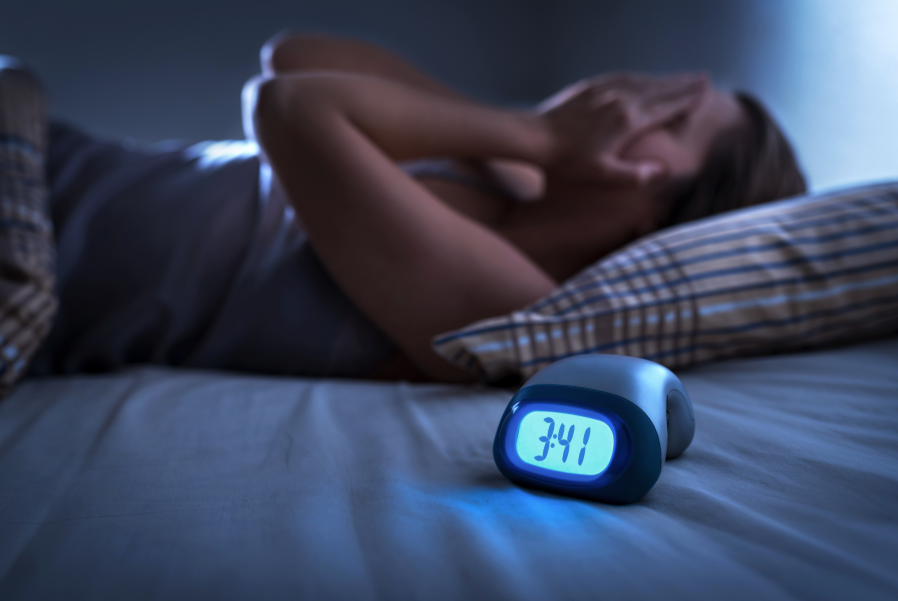 Woman laying in bed holding head in hands with alarm clock in the foreground